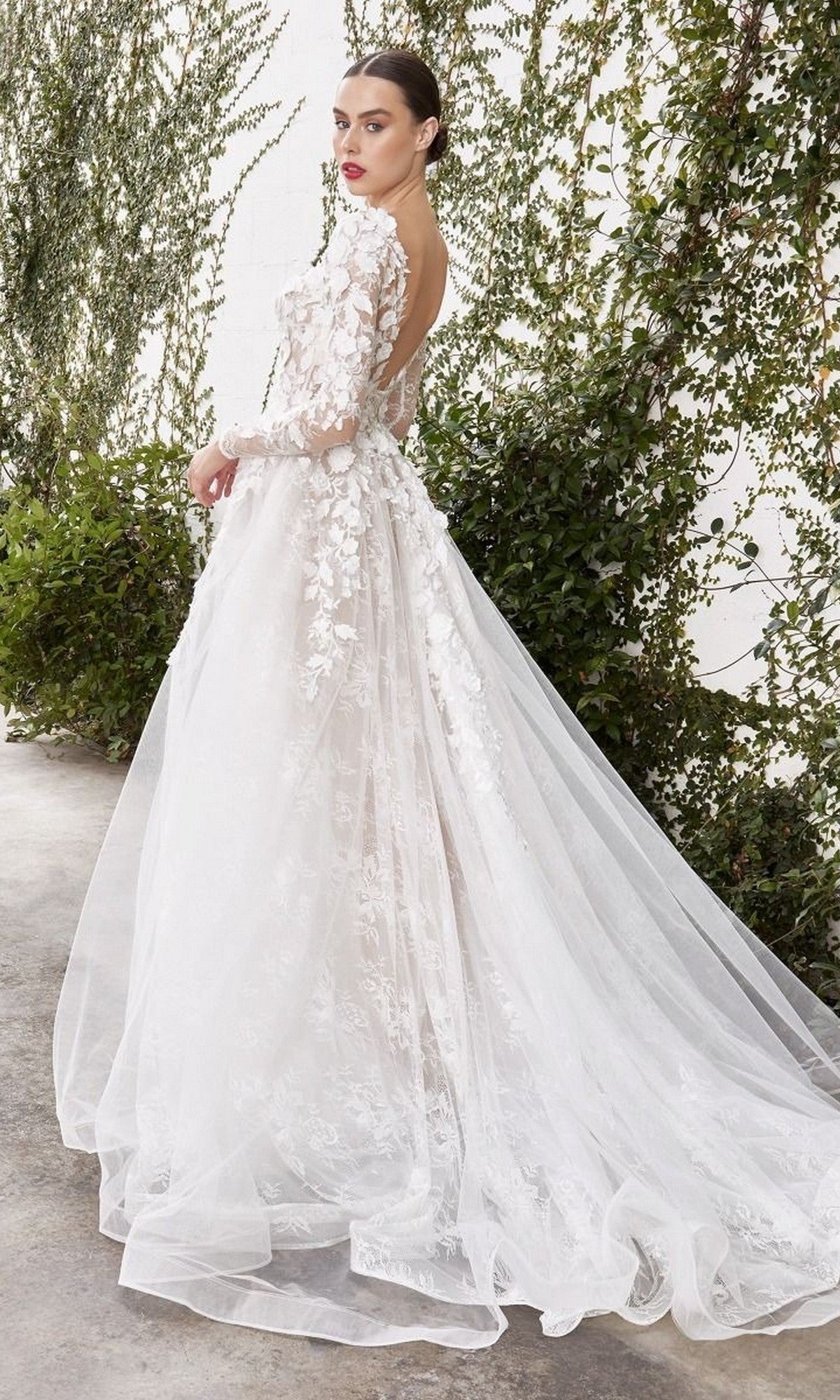 Wearing the Andrea and Leo A1067WC Long Formal Dress—a white gown with long sleeves, delicate floral lace appliques, and a sheer back—a woman stands before a green ivy-covered white wall, glancing over her shoulder.